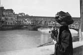 Ester Ellqvist-Bauer, med blommor i handen, står och tittar bort mot bron Ponte Veccio över floden Arno i Florens. I ett av husen till vänster, bodde de vid sitt besök i Italien.