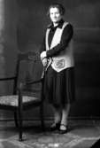 Ateljébild på en kvinna med stol och väst. Enligt Walter Olsons journal är bilden beställd av fröken J Bylund.