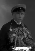 Ateljébild på en man som har tagit realexamen. Han har en blombukett och mössa. Enligt Walter Olsons journal är bilden beställd av Nils Johnsson.