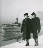 Två modeller i minkpäls och hatt på Södra Blasieholmskajen i Stockholm. Kungliga Operan och en skärgårdsbåt i bakgrunden.