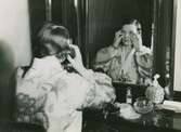 Kvinna i morgonrock gör ansiktsbehandling vid en spegel.
