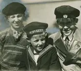 Skolungdom, pojken längst till vänster i tröja, skjorta, slips och basker, pojken i mitten i sjömanskostym och skärmmössa, pojken till höger i skjorta, slips, kavaj och skärmmössa.