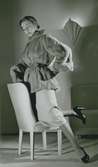 Modell i pälsjacka med skärp, pennkjol och pumps, på en stol.
