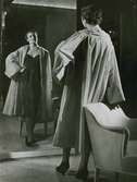Modell i mörk klänning och karamell-skär aftonkappa i faille, speglar sig framför en fåtölj. Från Edward Molyneux för Idun.