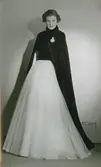 Modell i fotsid klänning med vitt tyll och hellång cape med brosch. Efter Balenciaga.
