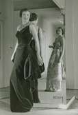 Modell i aftonklänning i sammet och pälshalsband tittar i en spegel, modell i mönstrad aftonklänning i bakgrunden. Av Maggy Rouff.