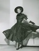 Modell i klänning med fluga, hatt, handskar och pumps.