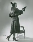Modell i pälskappa, hatt med rosett, handskar och pumps.