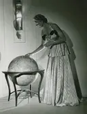 Modell i aftonklänning med volang och rosetter, tittar på en jordglob.