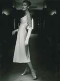 Modell i vit klänning, aftonhandskar, skor med stilettklack och örhängen i en mörk gång, NK:s Franska damskrädderi.