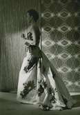 Modell i aftonklänning med blommigt mönster och rosett. Från Patou. Textilier i bakgrunden.