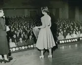 Tonårsmodell framför publik, kjol, blus och pumps.