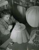Nordiska Kompaniet. En kvinna i arbetsrock monterar ett band på en lampskärm. På bordet bredvid henne, en sax och trådrullar. Text med blyerts på baksidan: 