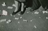 Mans- och kvinnofötter på ett golv med pappersskräp och apelsinskal. Text med blyerts på baksidan: 