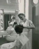 Damfrisering. En kvinna blir kammad. Nordiska Kompaniet. Text med blyerts på baksidan: 