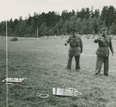Två uniformerade män på ett fält kastar ringar mot en träplatta med fyra pinnar. Nordiska Kompaniet. Text med blyerts på baksidan: 
