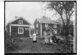 Tvåvånings enkelstuga med veranda med snickarglädje. Flygelbyggnad innehållande lanthandel, 4 personer framför byggnaden.
Konrad Eriksson