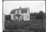 En och en halvplans bostadshus med veranda och frontespis, 10 personer framför huset.
Karl Linden