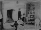 John och Ester Bauer på Björkudden utanför Gränna. Ester står innanför dörren och John sitter i en gungstol vid öppna spisen och läser tidningen.