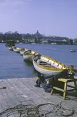 Träbåtar på rad vid kajen på Galärvarvet. Vy mot Skeppsholmen. Vilopaus i solen.