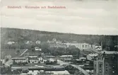 Vy över Norrmalm. Gamla gångbron (kallad Puckelbron och Kamelryggen) över Selångersån. Bron raserades vid vårfloden 1919. Lasarettet och stadsbacken. Hydéns möbelaffär ses till vänster efter Skepparegatan. Ett av varumagasinen i nedre höger kant. Vykort.