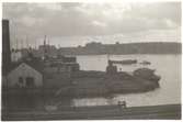 Vy över Beckholmen 1920-tal; varvsbyggnader, arbetsbåt och dockor samt bogserbåt med pråmar på Saltsjön.