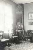 En salong i Karl Johansstil, med inkapslat ur, hörnplacerad spegel, stolar och en emmafåtölj.