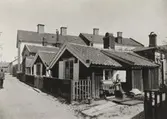 Små bostadshus med fönsterluckor och träfasader på Västerlånggatan i Gamla staden år 1908.