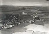 Flygfoto över Sandby. Byn med hus, ladugård, ekonomibyggnader och kyrkan samt grusvägen genom byn och åkrar.