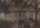 En man och två kvinnor i trädgården utanför ett hus med glasveranda.