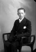 Ateljébild på en man i kavaj som sitter på en pall. Enligt Walter Olsons journal är bilden beställd av Arne Nilsson.