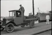 Bil med flak skjutsar manskap, t h officer; i bakgrunden örlogsfartyg vid kajkanten.
