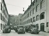 Parkerade Volvo-lastbilar med Nordiska Kompaniets varumärke på dörrarna. Text med blyerts på baksidan: NK:s nya lagerhus invid Stockholms stads slakthus i Enskede. 'Centrallager'. Sept. 1940