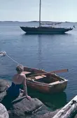 Kvinna med baddräkt på klipphäll i skärgården, segelbåten Elsi-Anne och en mindre roddbåt ligger förtöjda i närheten.