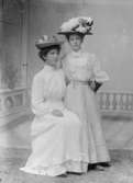 Ateljébild av två unga damer klädda i vita långklänningar och hattar.