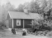 Liten stuga, herre och dam sittande framför. 

Fotograf: Ellen Kock? Axel Lundbäck m.fl.
Gullspångssamlingen.
Bilder från Gullspång med omnejd.