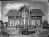 Exteriör. Trähus med flaggstång, fem herrar och två damer. Bilden otydlig. Gullspångssamlingen.Bilder från Gullspång med omnejd.