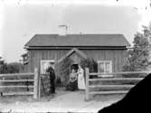 Exteriör.
Två damer och en herre framför liten brädfodrad stuga. 
Gullspångssamlingen. 
Bilder från Gullspång med omnejd.