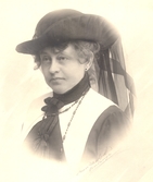 Bröstbild av Ellen Warholm i stor svart hatt med på sidanuppvikt brätte och långt svart flor bak, samt vit krage med midjelånga snibbar.