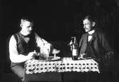 Två män som dricker snaps vid ett bord. Möjligen kan det vara ett trickfoto, där fotografen Karl Johan Östergren dubbelexponerat samma person.