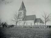 Exteriörbild av Havdhems kyrka på Gotland taget före 1912 (då tornets gallerivåning togs ned). Ett hästekipage med två män i förgrunden. Framför kyrkmuren står en staketliknande anordning hästarna förmodligen bands vid under kyrkobesöket.