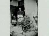 En äldre kvinna med spinnrock sitter på en stengård vid ett hus. I dörröppningen bakom henne står en kvinna och en liten flicka.