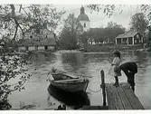 Flicka och pojke på brygga med båt. På andra sidan vattnet en kyrka, ett timrat hus samt en stor stenbyggnad.