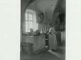 Interiörbild av Tjärby gamla kyrka. Fyra kvinnor sitter i kyrkbänkarna och en står. Severin Nilsson dokumenterade den innan den revs 1906.