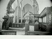 Interiörbild av okänd kyrka, rikt dekorerad. Vy över långskeppet med slanka pelare mot orgelläktaren. Till vänster i bild predikstol med genombruten baldakin och till höger korbänkar och psalmnummertavla.