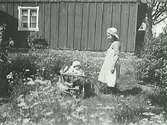 Två barn i en blomstrande trädgård med bostadshusets gavel i bakgrunden. Babyn sitter i en sorts kärra med en flicka intill sig.