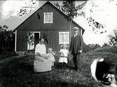 Familjebild. Ett par med två barn i trädgården, med späda träd, framför en husgavel (se även E5362).