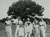 Fyra kvinnor och fyra barn, varav en pojke med stor keps, uppställda på ett fält framför ett stort träd. Kvinnorna har slipsar, troligen lärarinnor.