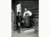 Man och kvinna i folkdräkt, troligen Daladräkt från Rättvik. Han sitter på en trappa till ett timrat hus och hon står bredvid.
