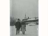 Två barn står i snön, flickan håller i böcker. I bakgrunden står ett timrat hus med vedtravar och en eka framför. Mariefreds kyrkspira sticker upp över taket. (Samma motiv som E5418)